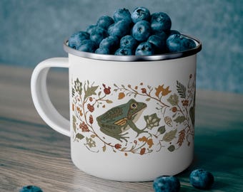 May include: A white enamel mug featuring a graphic illustration of a green frog surrounded by a wreath of brown and green autumn leaves, berries, and vines. The mug is filled to the brim with fresh, dark blue blueberries, with more scattered nearby.