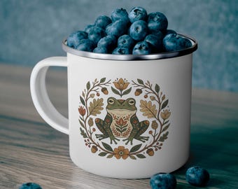 May include: A white enamel mug with a silver rim, filled with fresh blueberries. The front of the mug features a detailed illustration of a green frog centered within a circular wreath of orange flowers, brown acorns, and tan oak leaves.