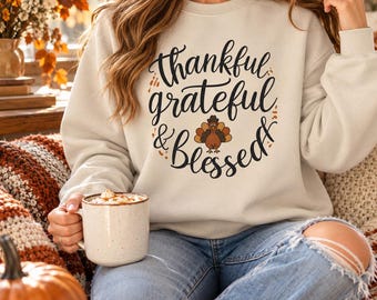 May include: A cream-colored crewneck sweatshirt featuring black cursive text that reads thankful grateful & Blessed, with a small graphic of a brown turkey centered below the text. The sweatshirt has long sleeves and a ribbed neckline.