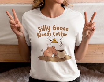 Op de afbeelding: Wit T-shirt met de tekst "Silly Goose Needs Coffee" in bruine letters. De afbeelding toont een cartoon gans met een trui, zittend aan een tafel met een koffiemok. De gans heeft de ogen dicht en er komt stoom uit de koffie.