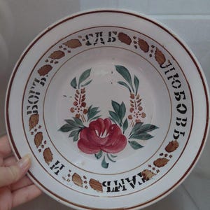 May include: White ceramic plate with a red rose and green leaves in the center. The rim features brown and black text and decorative elements. The plate has a brown border.