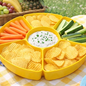 Può includere: Un vassoio da portata giallo diviso pieno di patatine, bastoncini di carote e fette di cetriolo, con una salsa al centro. Fragole in una tazza rossa a pois e un cestino da picnic con frutta sullo sfondo.