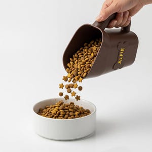 Peut inclure: Une pelle à nourriture pour animaux de compagnie marron, avec le nom "ALFIE" en jaune, verse des croquettes dans un bol en céramique blanc. La nourriture contient des morceaux en forme d'étoile. La pelle a une poignée.