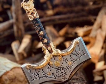 May include: A detailed, silver-colored hammer with gold accents and intricate engravings. The handle is wrapped in brown and black leather with gold detailing. The hammer's head features a decorative design and is resting on a wooden surface.