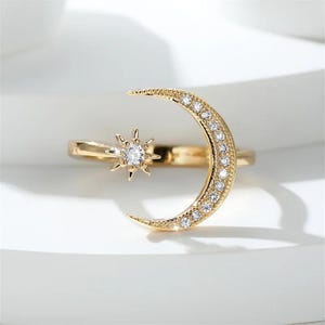 May include: A gold-colored ring featuring a crescent moon and a star design. The moon is encrusted with small, clear gemstones, and the star has a single, larger gemstone at its center. The ring is set against a white background.
