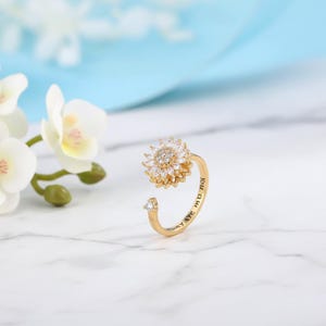 May include: A gold-colored ring with a sunflower design. The sunflower is made of small, clear crystals and has a central cluster of smaller crystals. The ring band has the inscription "You are my sunshine".