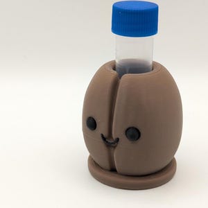Puede incluir: Un soporte marrón impreso en 3D con forma de grano de café con una cara sonriente, que sostiene un vial transparente con una tapa azul. El soporte tiene ojos negros y una boca curvada. El vial contiene un líquido oscuro.