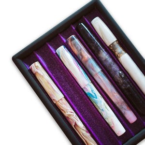 Könnte beinhalten: Sechs farbenfrohe Füllfederhalter mit Marmormuster in einer schwarzen Schale mit lila Samtfutter. Die Stifte weisen einzigartige Designs in Rosa, Blau, Weiß und Schwarz auf und sind in einer Reihe angeordnet.
