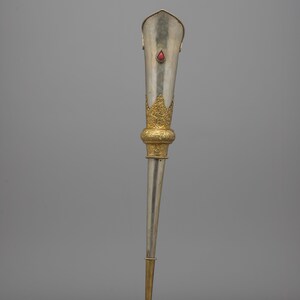 May include: A silver-colored ceremonial object with gold accents and a red gemstone. The top portion is flared and adorned with a gold filigree design. The base is gold, and the object tapers to a point.
