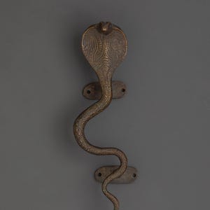 May include: A bronze-colored door handle shaped like a cobra snake. The handle features an intricately detailed cobra head and a winding body. The handle is mounted on a gray surface with two oval-shaped plates, each with two screw holes.