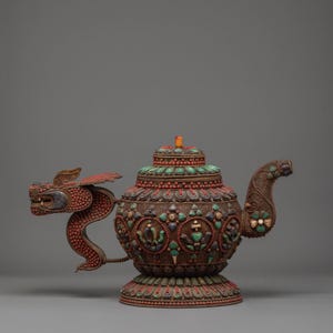 Może przedstawiać: Ozdobny brązowy czajnik z uchwytem i wylewką w kształcie smoka. Czajnik jest zdobiony czerwonymi, zielonymi i niebieskimi kamieniami. Pokrywka ma mały pomarańczowy kamień. Podstawa jest okrągła i ma szczegółowy wzór.