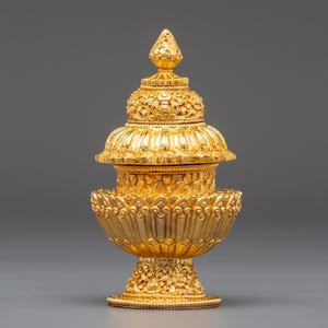 May include: A detailed, golden decorative object with intricate floral patterns. The object has a tiered design, with a base, a central section, and a pointed top. The gold color and ornate details suggest a ceremonial or religious item.