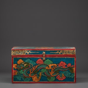 Puede incluir: Cofre rectangular de madera pintado de color turquesa con un diseño de dragón. El dragón es verde con detalles en naranja, sobre nubes naranjas y verdes. Borde rojo y cierre de latón.