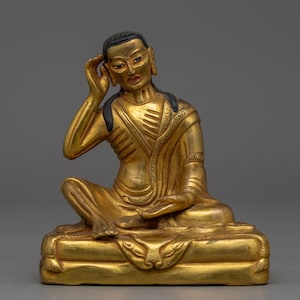 May include: A golden statue of a seated figure with one hand raised to the ear. The figure is wearing a draped garment and is seated on a decorative base. The statue has black hair and blue eyes.