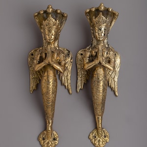 May include: Two brass door handles featuring a detailed design of a figure with a cobra headdress, wings, and a fish tail. The figures have their hands together in a prayer position. The handles are a golden color.