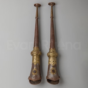 May include: Two long, slender, trumpet-like instruments made of dark brown metal with ornate gold-colored metal accents. Each piece features a flared base with a circular floral emblem, a decorative filigree band, and a narrow, elongated neck.