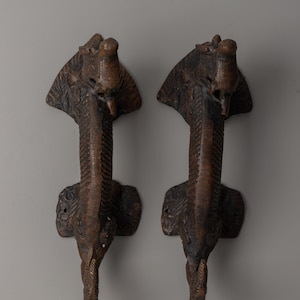 May include: Two bronze-colored, cobra-shaped door handles. Each handle features a detailed cobra head with an open hood and a textured body. The handles are mounted on a decorative base, showcasing intricate carvings.