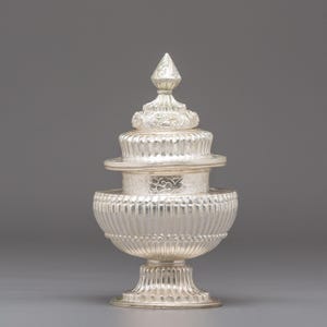 May include: A silver-colored, ornate decorative object with a tiered design. The object features a base, a ribbed central section, and a multi-layered top with a pointed finial. The surface has intricate detailing and a polished finish.