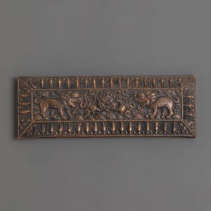 May include: A rectangular, bronze-colored relief carving with intricate details. The artwork features two elephant-like creatures flanking a central design with floral and mythical elements. The border is decorated with vertical lines.