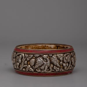 May include: A silver-colored bracelet with intricate carvings of birds and foliage. The bracelet features a band of red material around the top and bottom. The design is detailed and ornate, showcasing a vintage aesthetic.