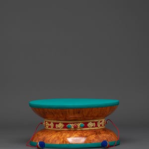 Puede incluir: Un tambor decorativo de madera con tapa y base turquesa. El tambor presenta un cuerpo marrón rojizo con una banda decorativa de acentos dorados, rojos y turquesas. Pequeñas borlas azules y rojas cuelgan de la parte inferior.