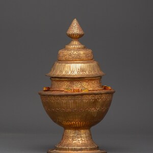 Puede incluir: Un recipiente dorado y ornamentado con un diseño escalonado. La base tiene forma de cuenco redondeado, sostenido por un pedestal. El recipiente presenta intrincados patrones y una parte superior cónica. Una pieza decorativa.
