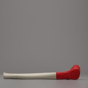 May include: A white and red object, possibly a decorative item or a prop, lies horizontally against a gray backdrop. The object has a long, cylindrical white section and a bulbous red end. The red section appears to be a solid, smooth material.