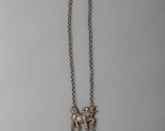 May include: Silver-toned necklace featuring a detailed pendant of a mythical creature with dangling charms. The necklace has a chain design and includes heart, bell, and leaf-shaped charms. The pendant is approximately 2 inches in length.