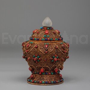 Puede incluir: Jarra ornamentada de estilo antiguo con un diseño de filigrana dorada. La jarra está adornada con piedras preciosas rojas, azules y verdes. Un remate transparente en forma de lágrima corona la tapa. La jarra tiene un patrón decorativo y detallado.