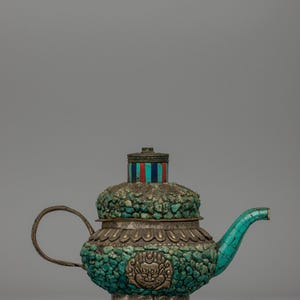 Puede incluir: Una tetera ornamentada con exterior de piedra turquesa y detalles plateados. La tetera presenta un asa, un pico y una tapa detallados con un diseño a rayas. Un emblema decorativo está en la parte delantera.
