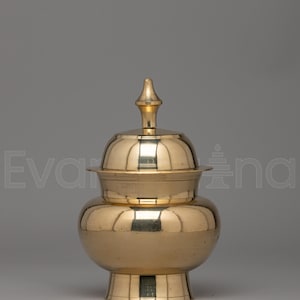 May include: A polished brass vessel with a rounded body, a flared base, and a removable lid featuring a pointed finial. The metallic surface has a reflective gold tone, and the container has a wide opening with a distinct rim below the lid.