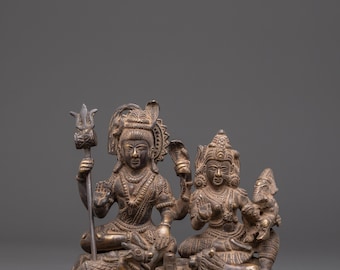 Puede incluir: Estatua de bronce que representa dos figuras sentadas, posiblemente deidades, con tocados y joyas ornamentadas. Una figura sostiene un tridente, la otra una serpiente. La estatua está sobre una base decorativa, mostrando detalles intrincados y artesanía.