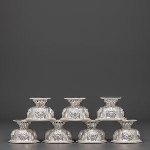 Pode incluir: Uma pilha de oito taças prateadas e ornamentadas com pés. Cada taça apresenta um design em relevo detalhado ao redor da base e um pequeno topo circular. As taças estão dispostas em forma de pirâmide sobre um fundo cinza escuro.