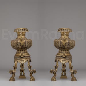 May include: Two antique brass urns with detailed ornamentation. Each urn has a rounded body, a decorative top, and is supported by three legs with animal figures. The brass has a warm, aged patina, suggesting a vintage origin.