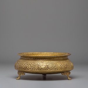 May include: A gold-colored, ornate bowl with a wide, shallow design. The bowl features intricate floral and geometric patterns around the sides and rests on three small, decorative feet. The rim has a textured, ridged design.