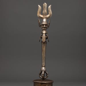 Puede incluir: Objeto ceremonial plateado en forma de tridente. La parte superior presenta tres púas sobre un diseño detallado de calavera. El eje es largo y delgado, con elementos decorativos. La base es una plataforma redonda y texturizada.