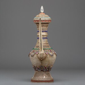 May include: An ornate silver-coloured pitcher with detailed patterns and colourful gemstones. The pitcher has a long spout and handle, with a decorative lid topped with a clear crystal. The base and body are decorated with red, blue, green, and purple stones.