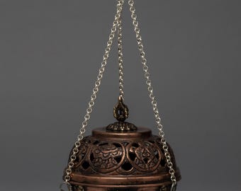 May include: A copper-colored, spherical incense burner with intricate cut-out designs and a decorative lid. It hangs from a silver chain. The burner features a detailed pattern and rests on three small feet. The overall design suggests a cultural or spiritual theme.