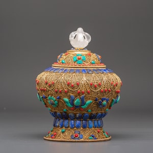 Peut inclure: Vaisseau doré orné d'un couvercle, orné de filigranes complexes et de pierres précieuses colorées. Le récipient présente des accents rouges, bleus et turquoise, et un fleuron clair et sculpté. La base et le couvercle sont décorés de motifs floraux et géométriques.