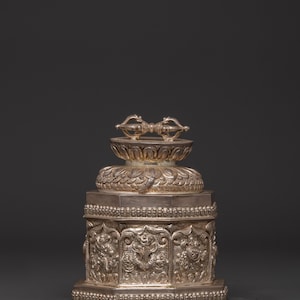 May include: A silver-colored, ornate religious artifact. The object features a detailed, multi-tiered design with intricate carvings and a symmetrical top element. The base is a geometric shape with detailed relief carvings.