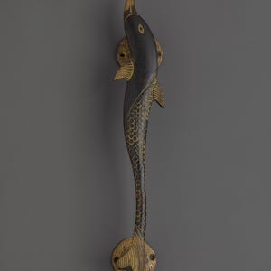 May include: A decorative door handle in the shape of a stylized fish. The handle is primarily black with gold accents, including the fins, tail, and a scale pattern along the body. The handle is mounted on a gray surface.