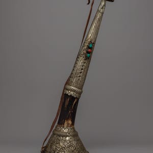 May include: A decorative horn instrument with silver-colored metal accents and ornate carvings. The horn features three colorful gemstones and a brown leather strap. The base of the horn is a dark brown color.