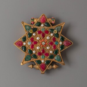 May include: A gold-coloured star-shaped pendant with a central floral design. The pendant is decorated with red, green, and clear gemstones, pearls, and a central diamond. It has a loop at the top for hanging.