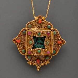 May include: A gold-colored pendant necklace with an ornate filigree design. The pendant features a central square with a carved teal figure, surrounded by emerald, ruby, and coral-colored gemstones. The necklace has a delicate gold chain.