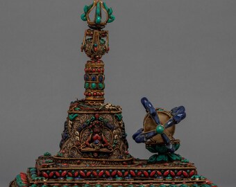 May include: An ornate, gold-colored religious artifact with intricate details. It features a central tower adorned with turquoise, red, and blue stones, and a smaller, multi-armed object. The base is decorated with similar stones and detailed carvings.