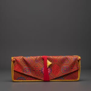 May include: A rectangular clutch bag with a patterned design in red, gold, and blue. The fabric features a geometric pattern. A red ribbon wraps around the centre, secured with a gold-coloured star-shaped clasp. The edges are trimmed with gold.