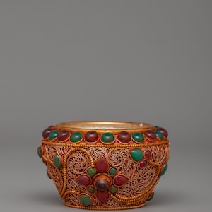 May include: Ornate gold-colored bowl with intricate filigree design. Adorned with red and green gemstones. The bowl has a wide rim and a detailed floral pattern, showcasing a blend of craftsmanship and elegance.