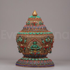Peut inclure: Un récipient décoratif en métal avec couvercle, présentant des motifs en filigrane argenté. La surface est ornée d'incrustations de turquoise, de corail et de lapis-lazuli, représentant une figure centrale assise et des motifs floraux.