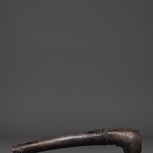 May include: A dark, metallic, curved object with ornate detailing. The object has a bulbous end and a flared opening, suggesting it may be a horn or a similar instrument. The surface shows signs of wear, indicating age.