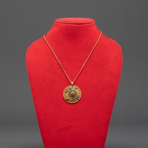 Può includere: Una collana con pendente dorato con un design circolare. Il pendente presenta un motivo dettagliato con pietre rosse e turchesi. La collana è esposta su un busto rosso su sfondo grigio.